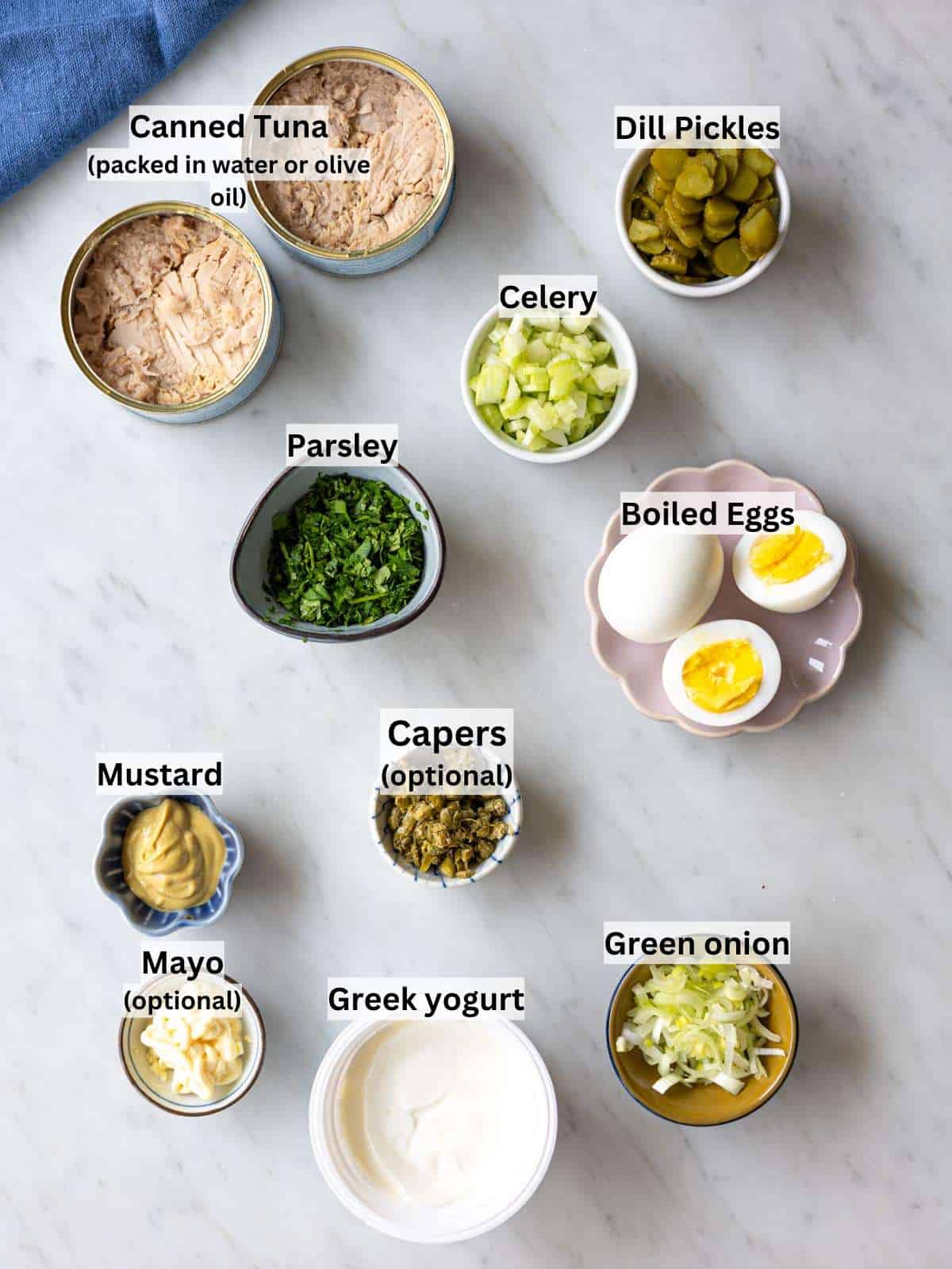 Ingredients for tuna egg salad arranged on a light counter with tuna, eggs, yogurt, herbs.