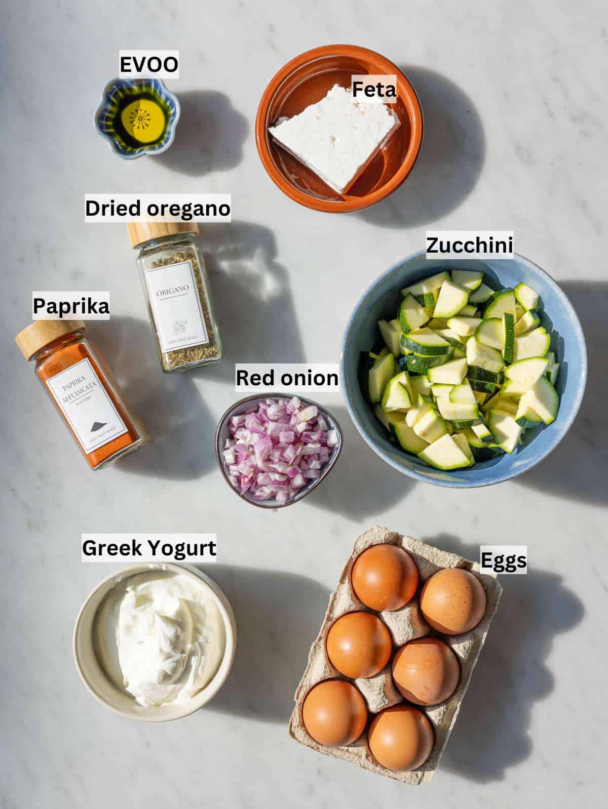 Fresh eggs, thick Greek yogurt, and chopped zucchini arranged on a bright marble surface in natural light.