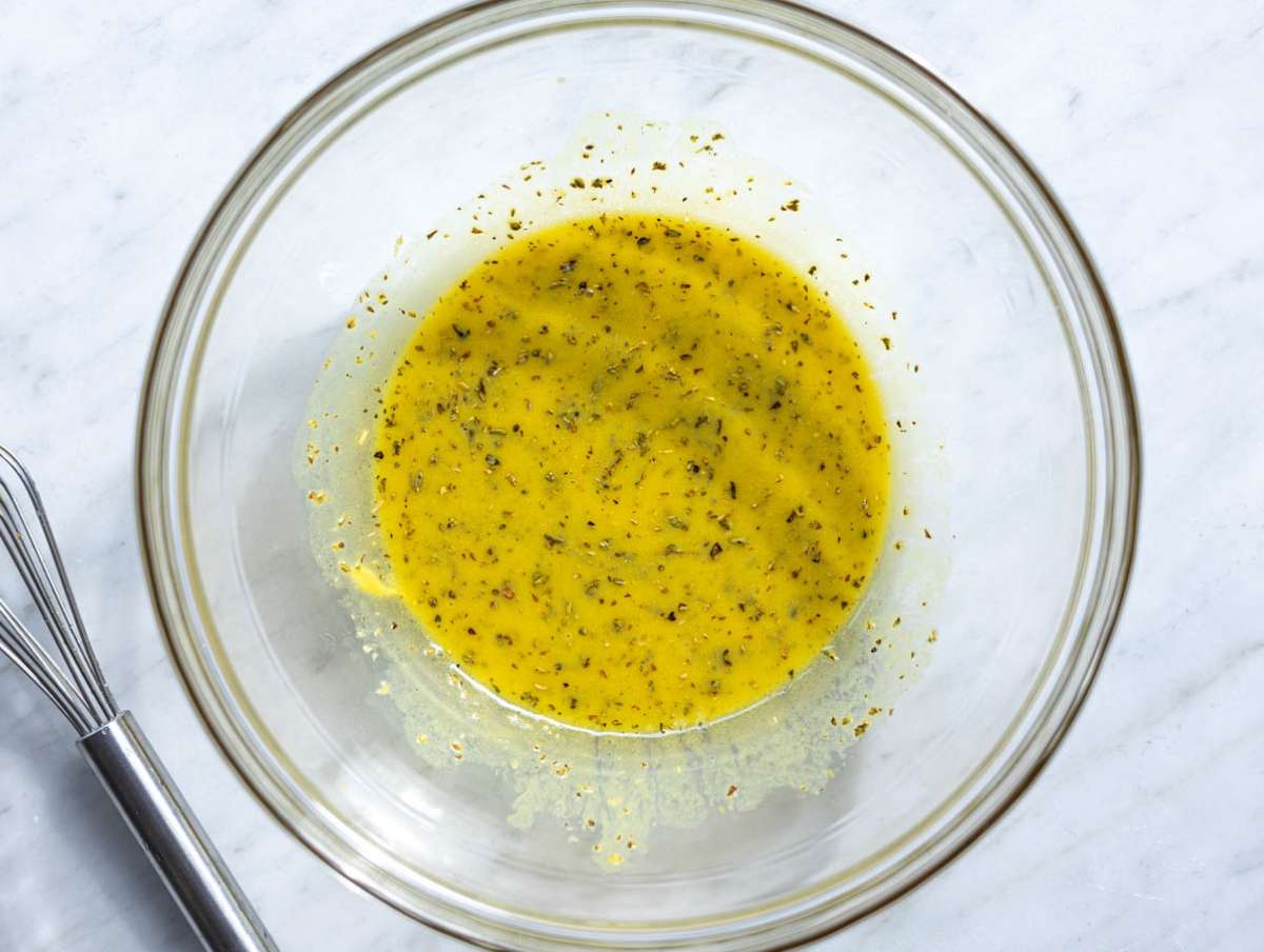 Step 1 Whisking lemon oregano dressing in glass bowl on marble table.