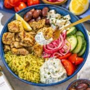 Mediterranean chicken bowl with lemon garlic chicken, yellow rice, tzatziki, fresh vegetables, and olives on marble surface.