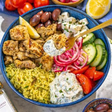Mediterranean chicken bowl with lemon garlic chicken, yellow rice, tzatziki, fresh vegetables, and olives on marble surface.