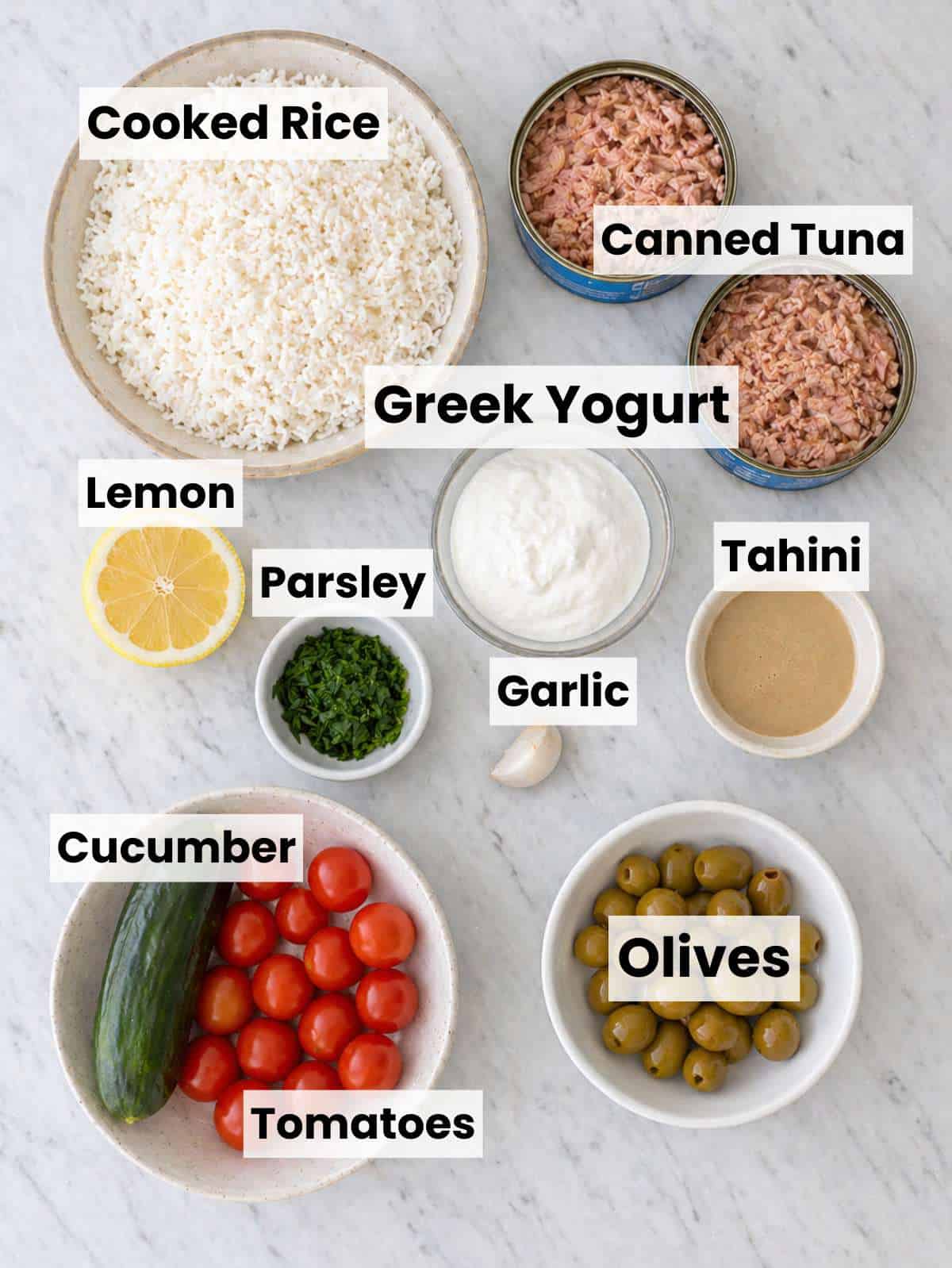 Tuna rice bowl with Ingredients arranged on marble surface with tuna, rice, yogurt, tahini, lemon, fresh vegetables tuna, cucumber, tomatoes, olives, and pickled onions on marble table.