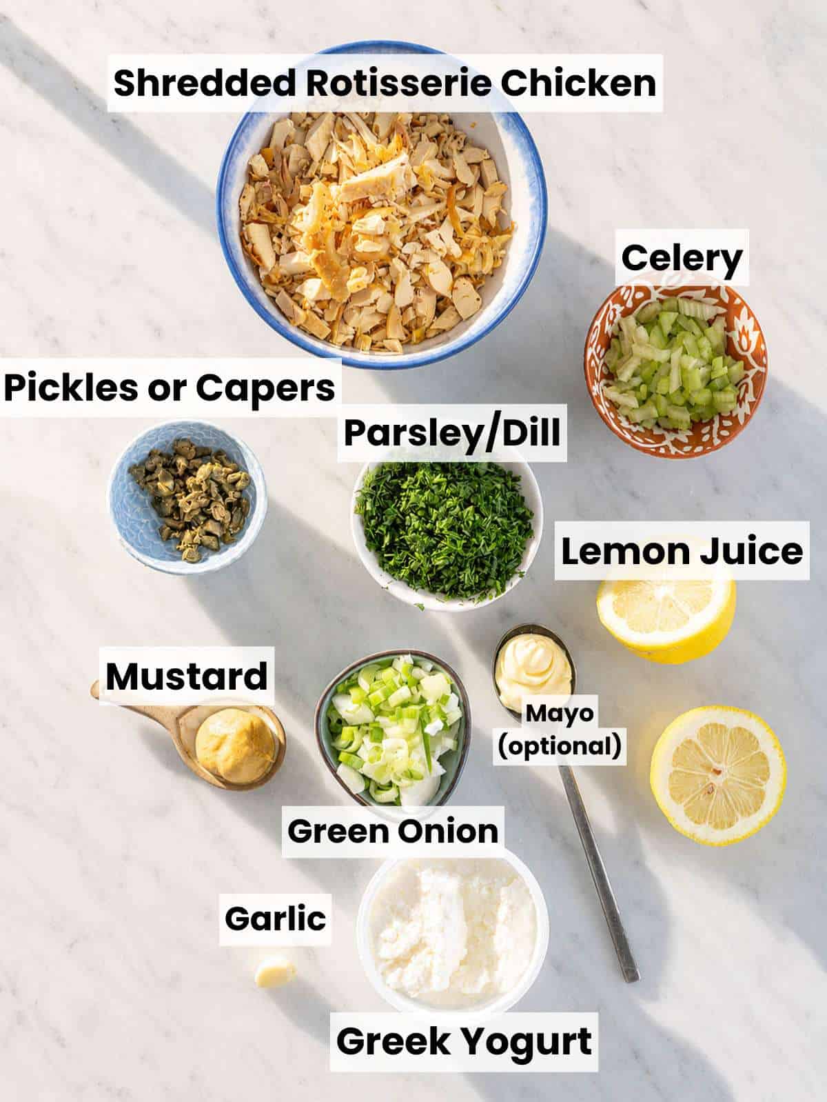 Shredded chicken, cottage cheese, celery, herbs, pickles, lemon, and Dijon mustard on marble counter.